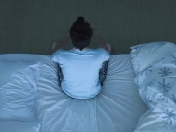 A man in a white shirt and grey sweat pants sits at the edge of a bed. 