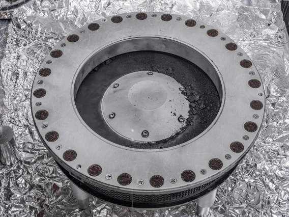 A round, metal canister with gray rocks and dust piled on the top