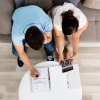 Overhead view of a man and woman sitting on a sofa, reviewing financial documents. The man is writing on a piece of paper in a binder while the woman uses a calculator, both appearing focused on their task.