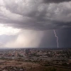 A large storm cloud hovers over a Southwestern metropolitan area. In the distance, rain falls as a bolt of lightning flashes.