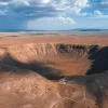 an aerial view of a meteor crater under cloudy blue skies on a vast desert landscape